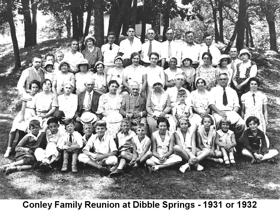 Black and white photo of several dozen men, women, and children standing or sitting in descending rows on a steep wooded hillside. A small frame building is visible behind them amongst the trees at the top of the hill. Sarah Dibble Conley, with white hair, is visible at the center of the crowd; behind her and to her right is Ora Conley Dreher.